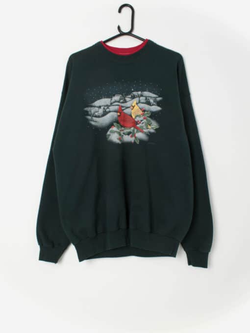 A sweatshirt on a hanger against a white wall. The sweatshirt shows graphics of a Christmas theme including Cardinal Birds, snow and a little town contrasted with a Forest Green background, and red trim crew neck.